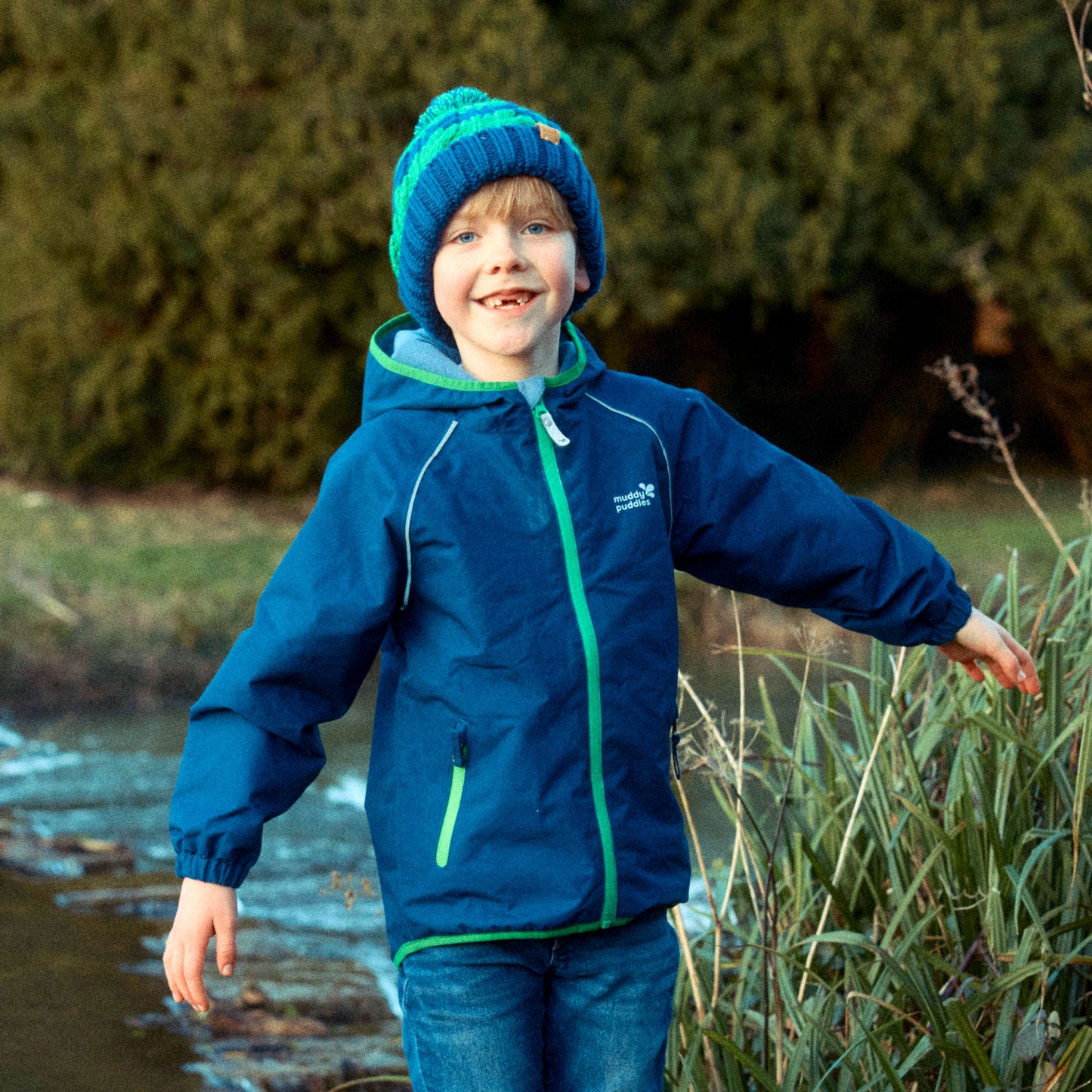 A happy, gap-toothed boy in a Navy EcoSplash Waterproof Fleece Lined Jacket is stood outside by some reeds and a stream.