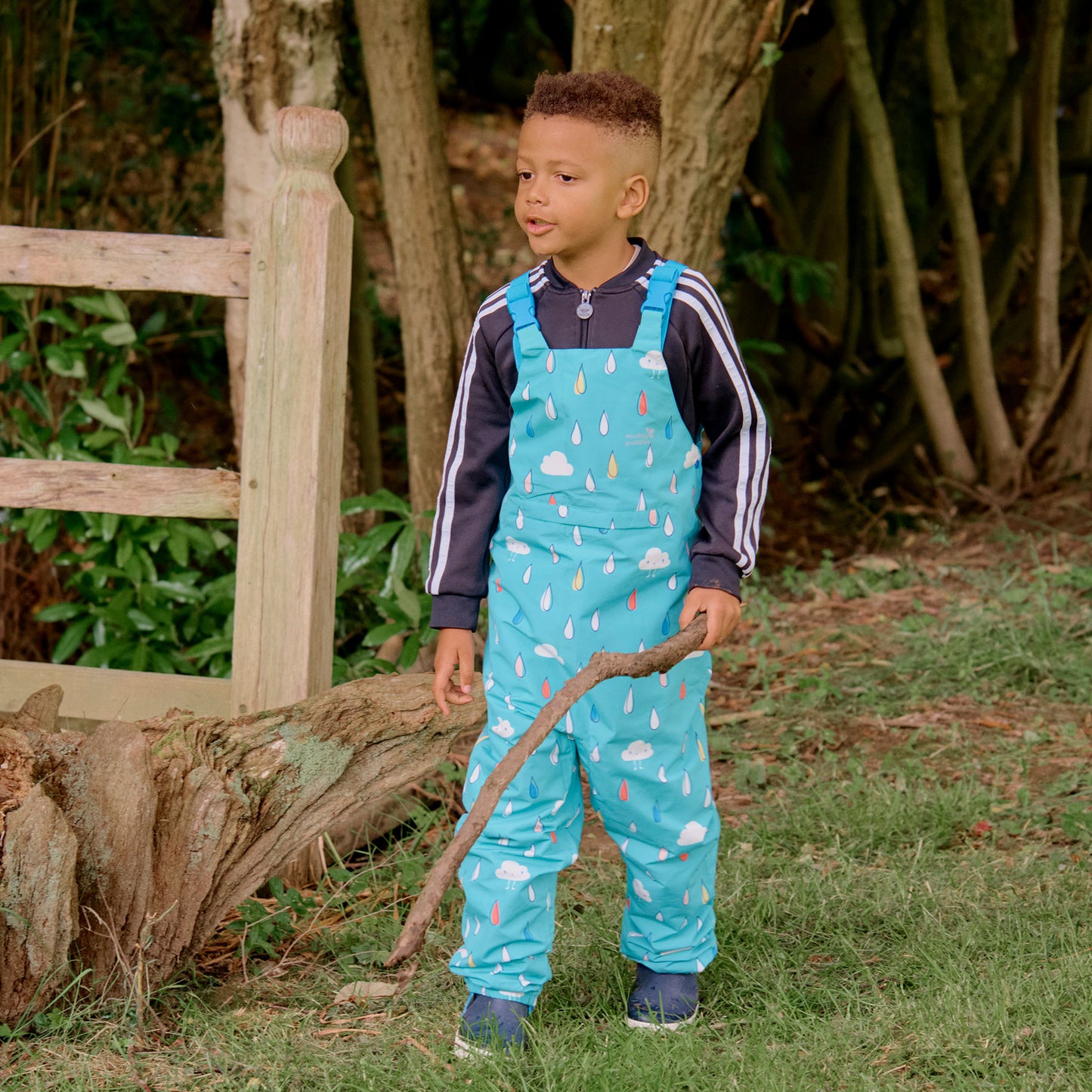 A boy in EcoLight Waterproof Dungarees Green Raindrops stands in a garden, holding a stick and looking to the side.