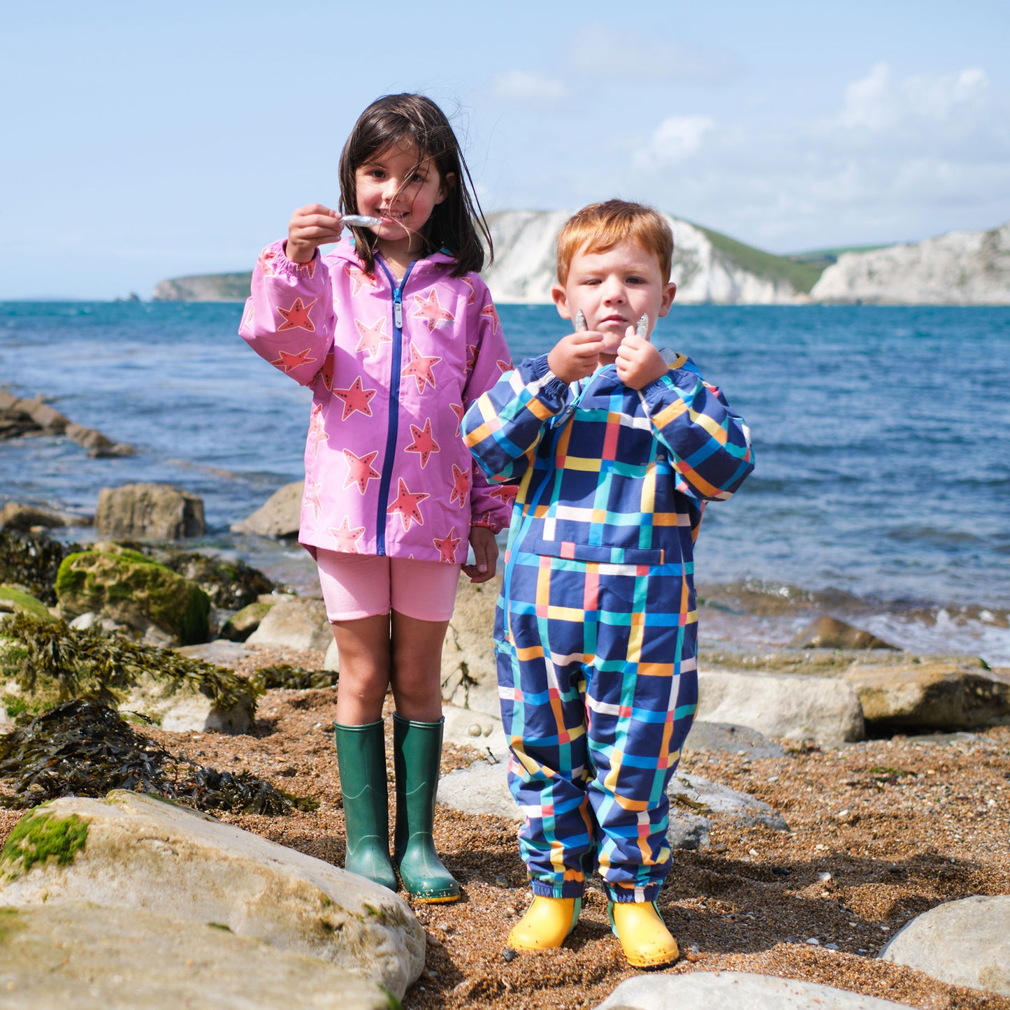 A girl and boy on a beach, holding fish, the boy wearing EcoLight Lightweight Waterproof Puddle Suit Multi Check.