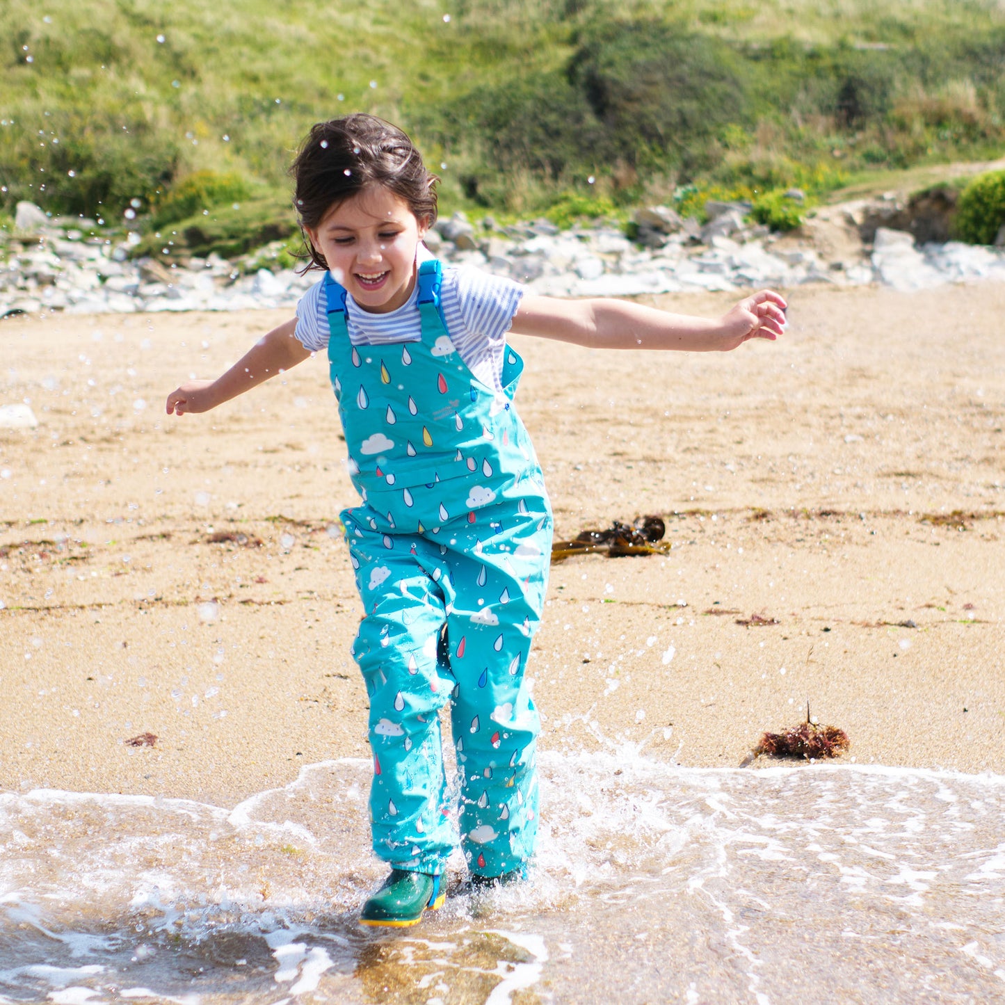 A young girl in wellies and blue dungarees with rain drop pattern playing on the beach, splashing in the sea with arms outstretched.