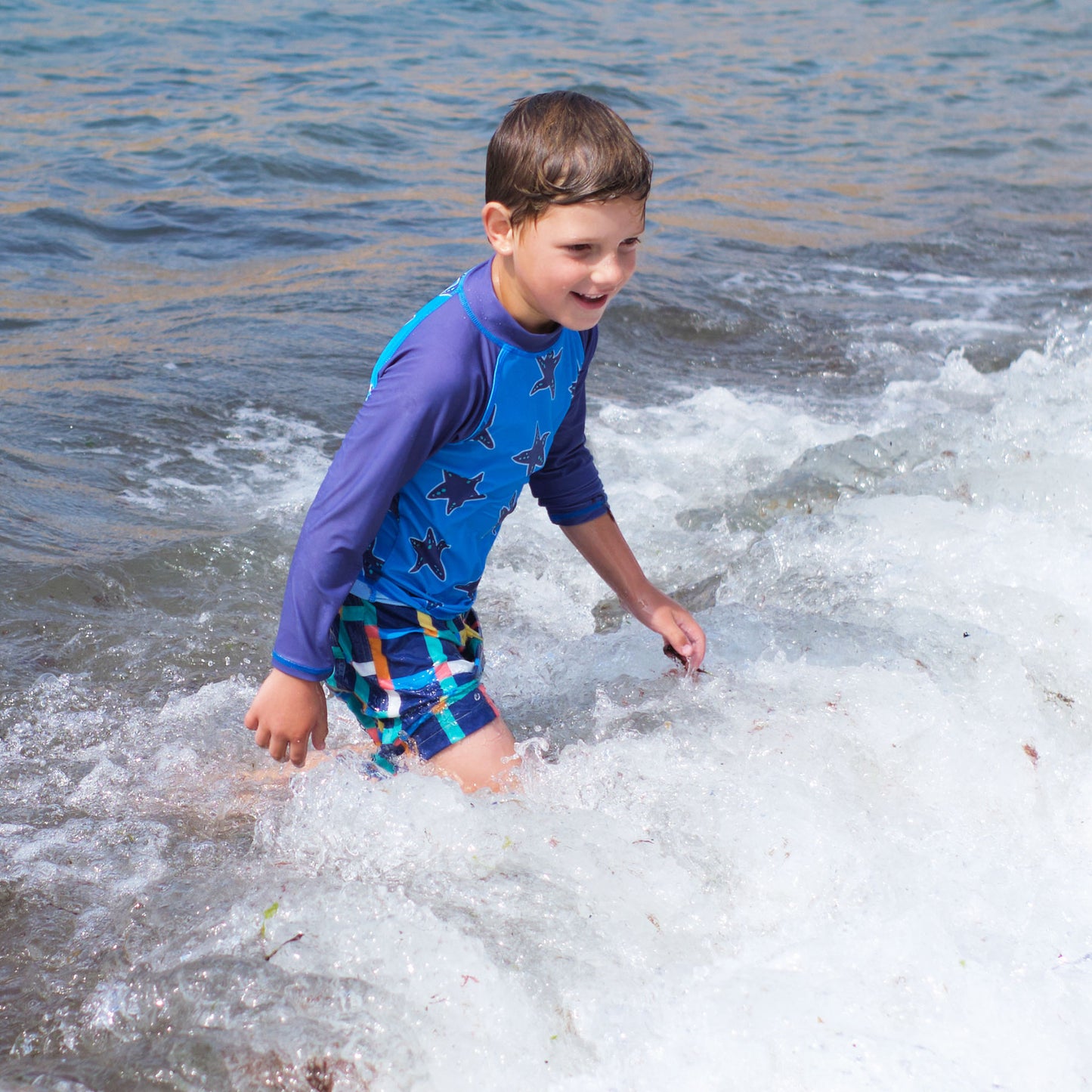 A young boy in a blue star-patterned rash vest and UV Protective Swim Shorts Multi Check, playing happily in the sea on a sunny day.