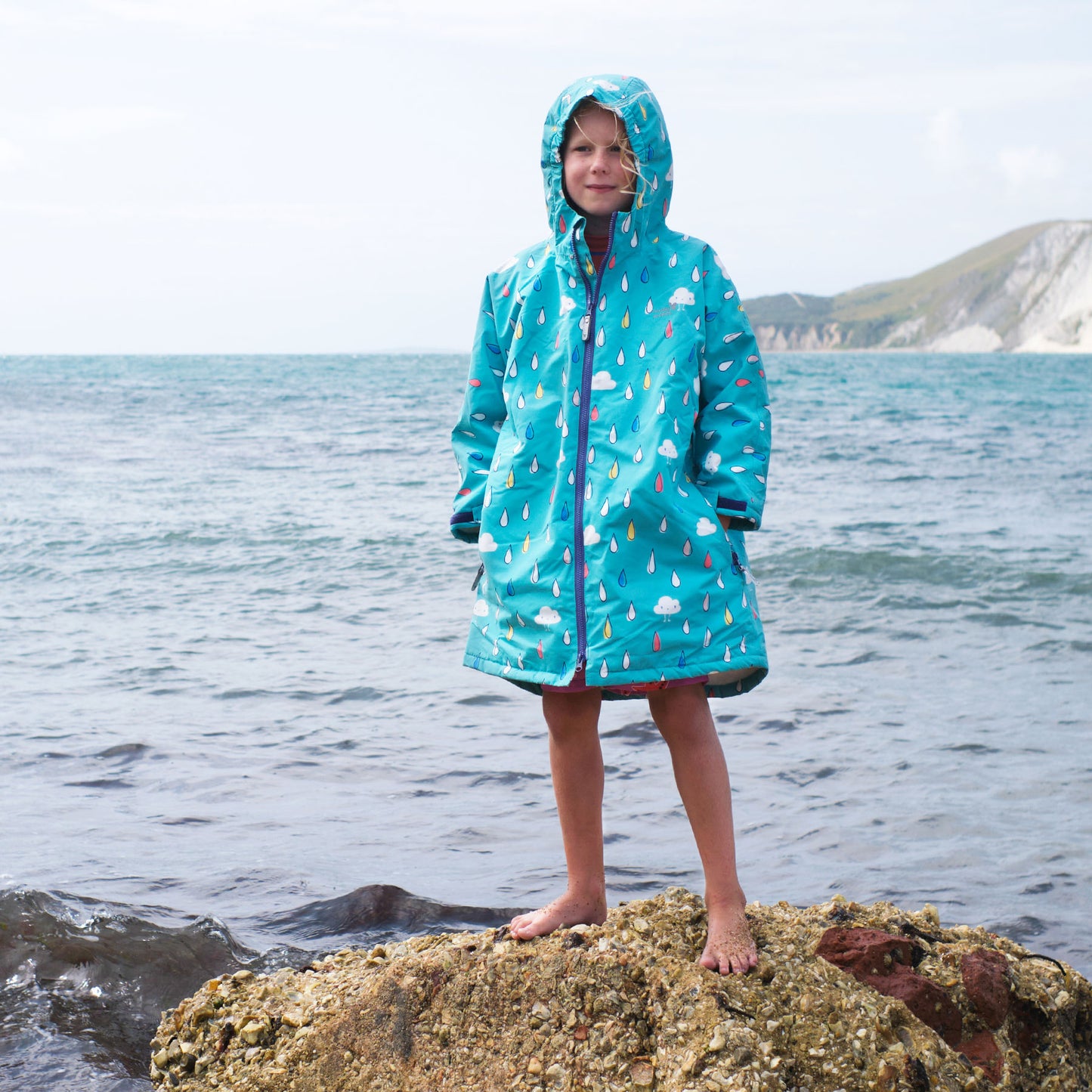 A girl on a rocky beach in a Waterproof Changing Robe Green Raindrops, standing on a rock with the sea and cliffs in the background.