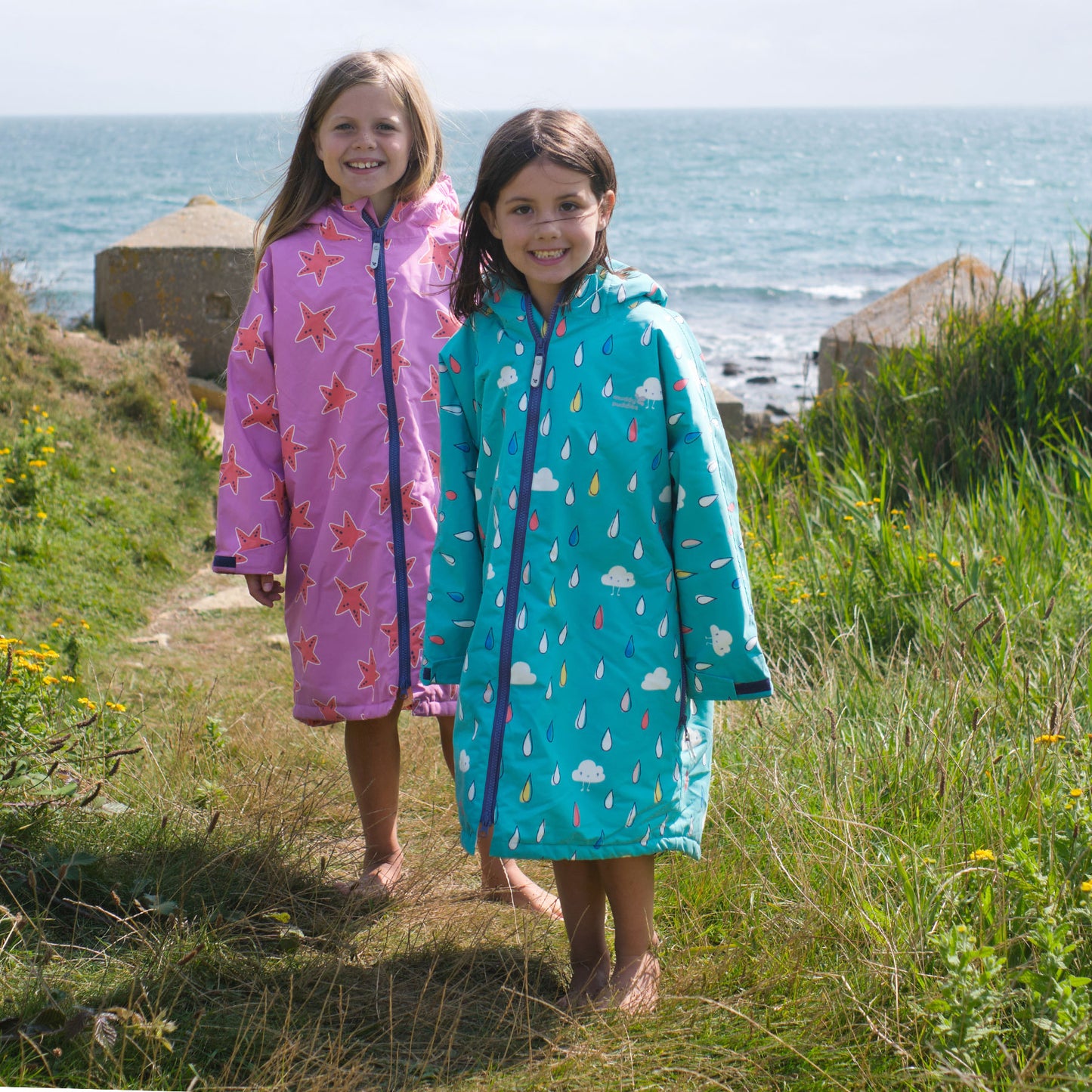 Two children wearing Waterproof Changing Robes. One is green raindrops and one is pink stars, posing near the sea in a grassy field.