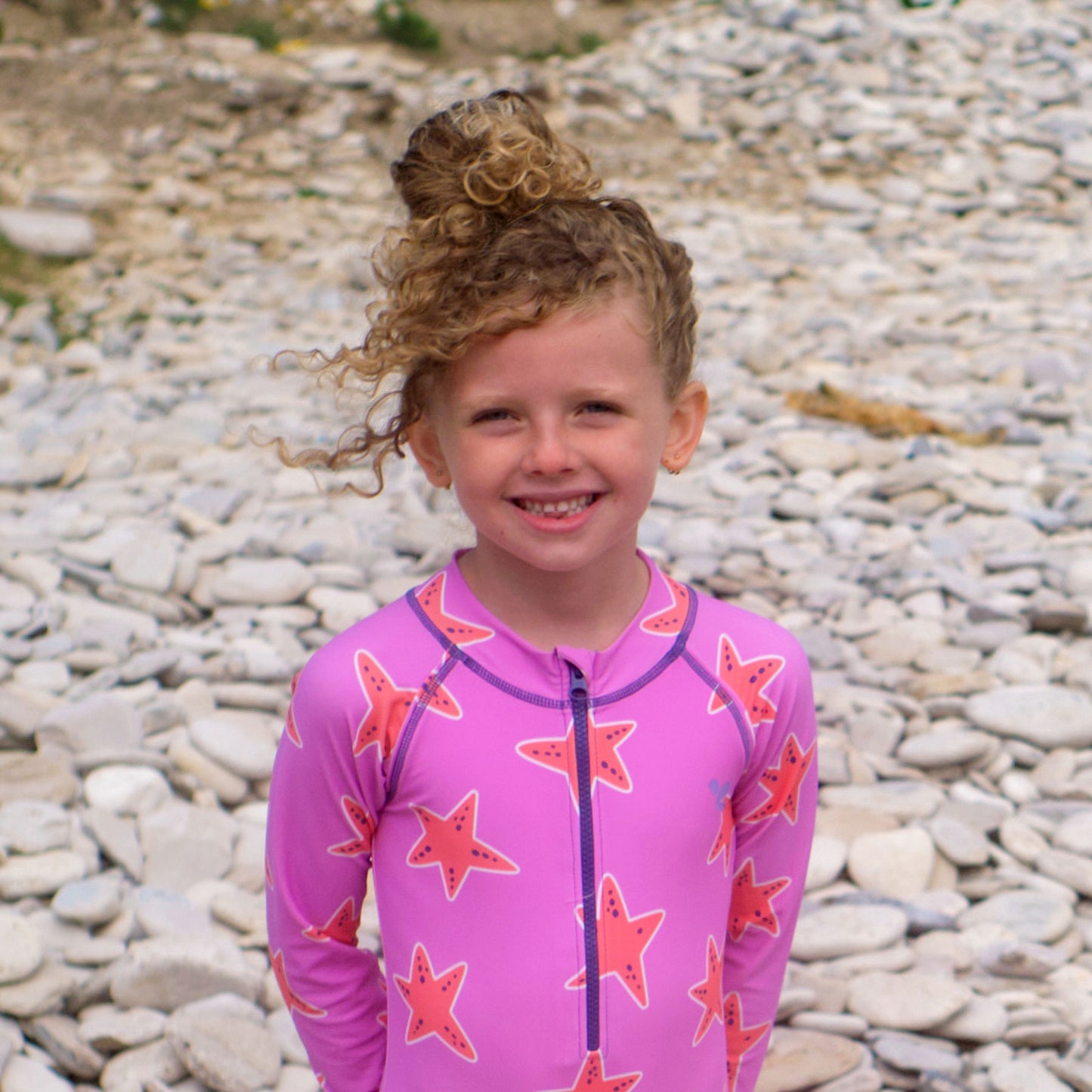 A smiling child in a UV Protective Long Sleeve Swimsuit Pink Starfish stands on a pebble beach, with curly hair in a bun.