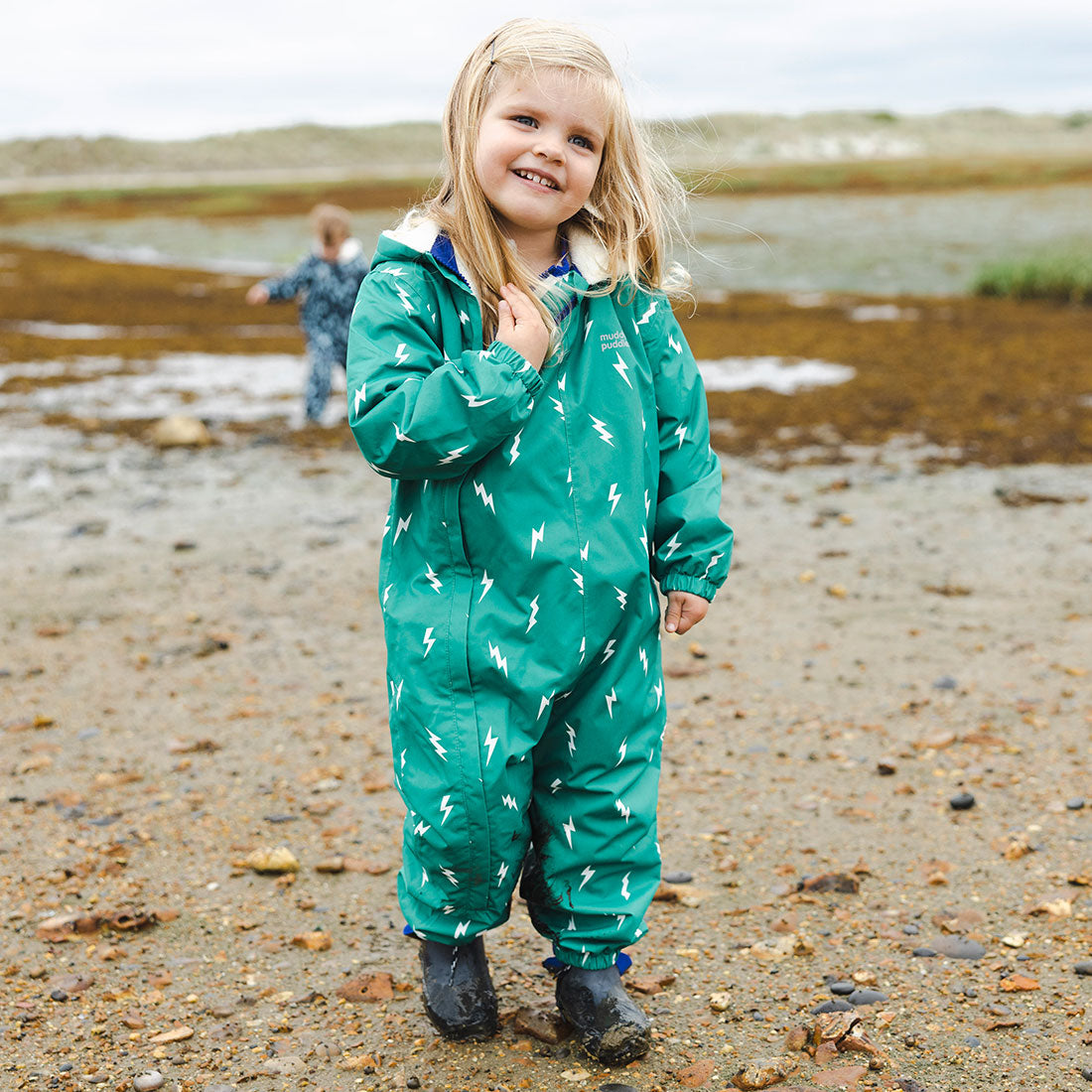 A young child wears a "3 in 1 Waterproof Scampsuit Green" patterned with lightning bolts, standing on a beach with another child in the background.