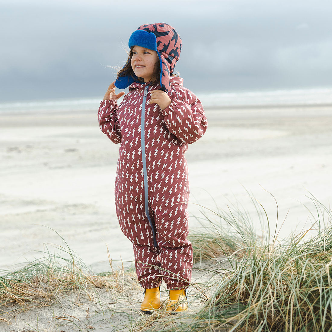 A child on a beach in an EcoSplash waterproof pink puddle suit with lightning bolts, wearing a hat and yellow wellies.