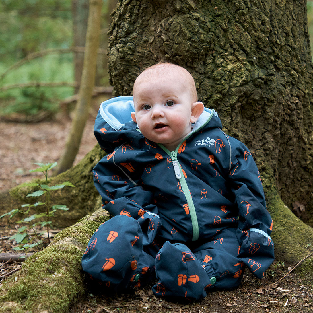 A baby sits by a tree wearing a Navy Print EcoSplash Waterproof Fleece Lined Puddle Suit in a forest, looking at the camera.