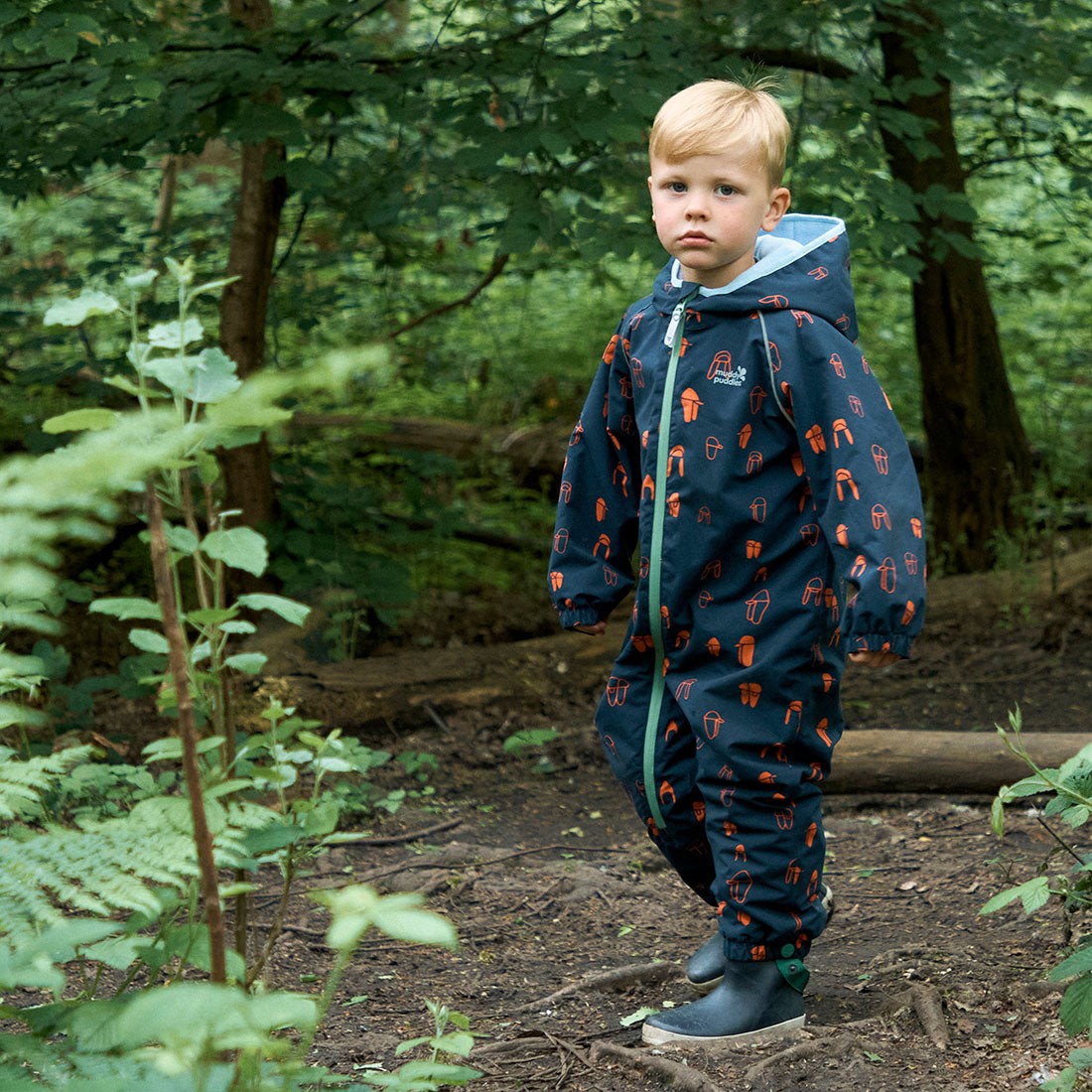 A young boy standing in a woodland scene wearing the EcoSplash Waterproof Fleece Lined Puddle Suit Navy Print.