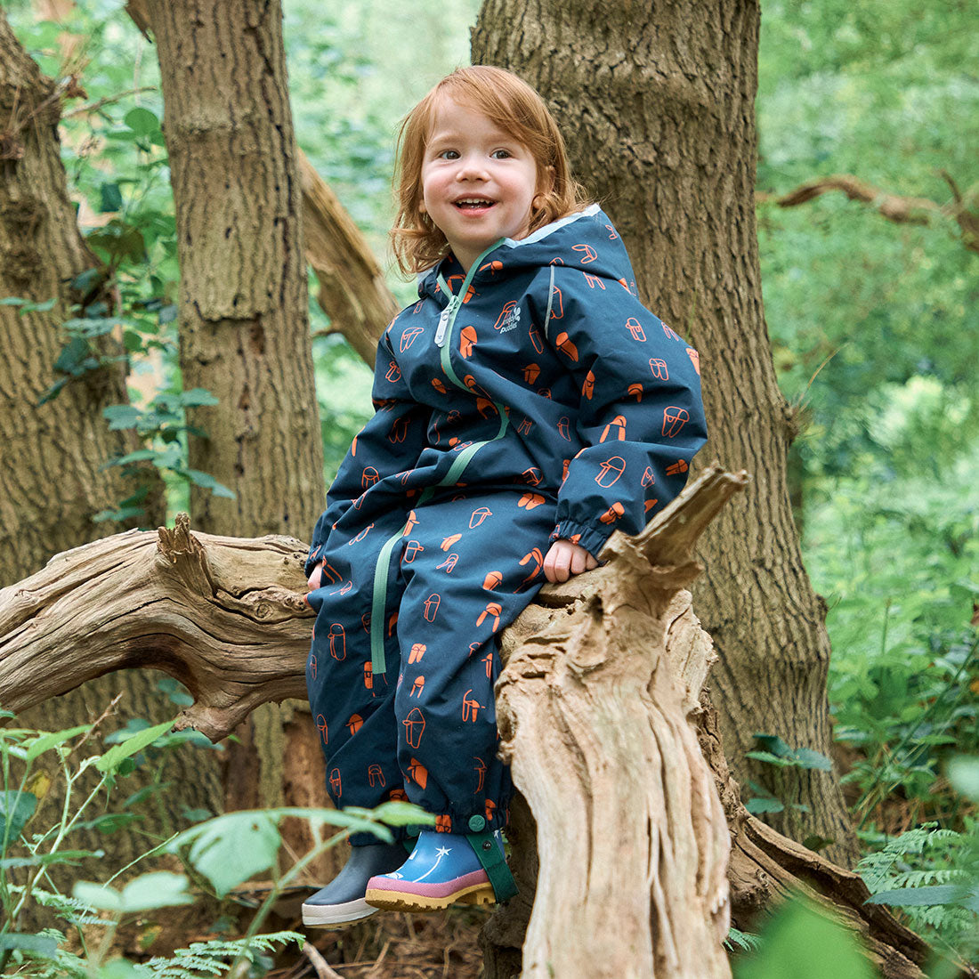 A child sits on a tree branch in a forest, wearing a Navy Print EcoSplash Waterproof Fleece Lined Puddle Suit and colourful wellies.