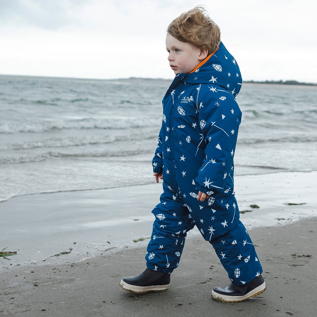 A young child is on a beach wearing a blue EcoWarm Waterproof Snowsuit Blue Sky patterned with stars and other shapes.