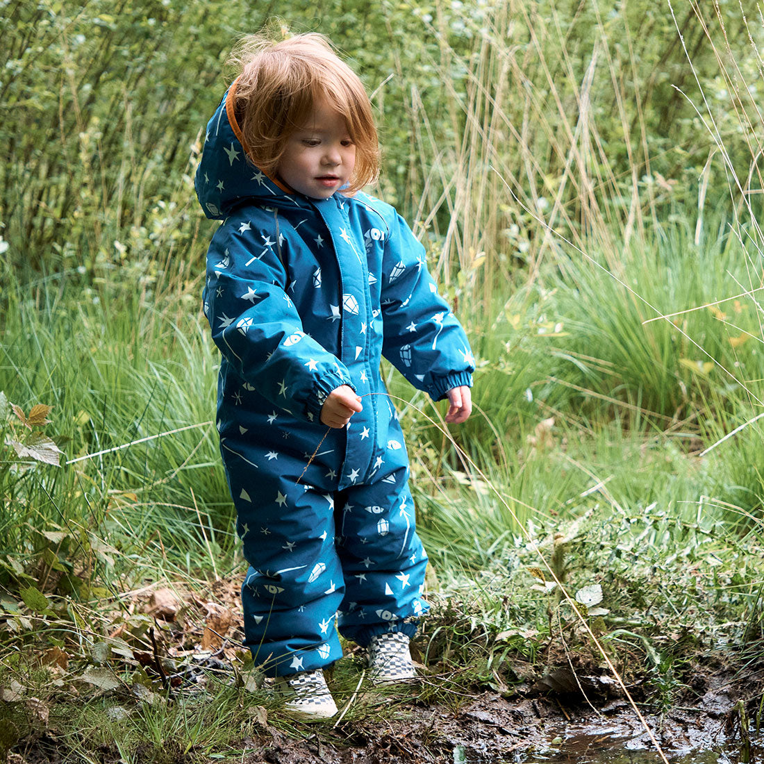 A toddler in a Blue Sky EcoWarm snowsuit explores a muddy patch in the woods. The snowsuit features a star and gem pattern.