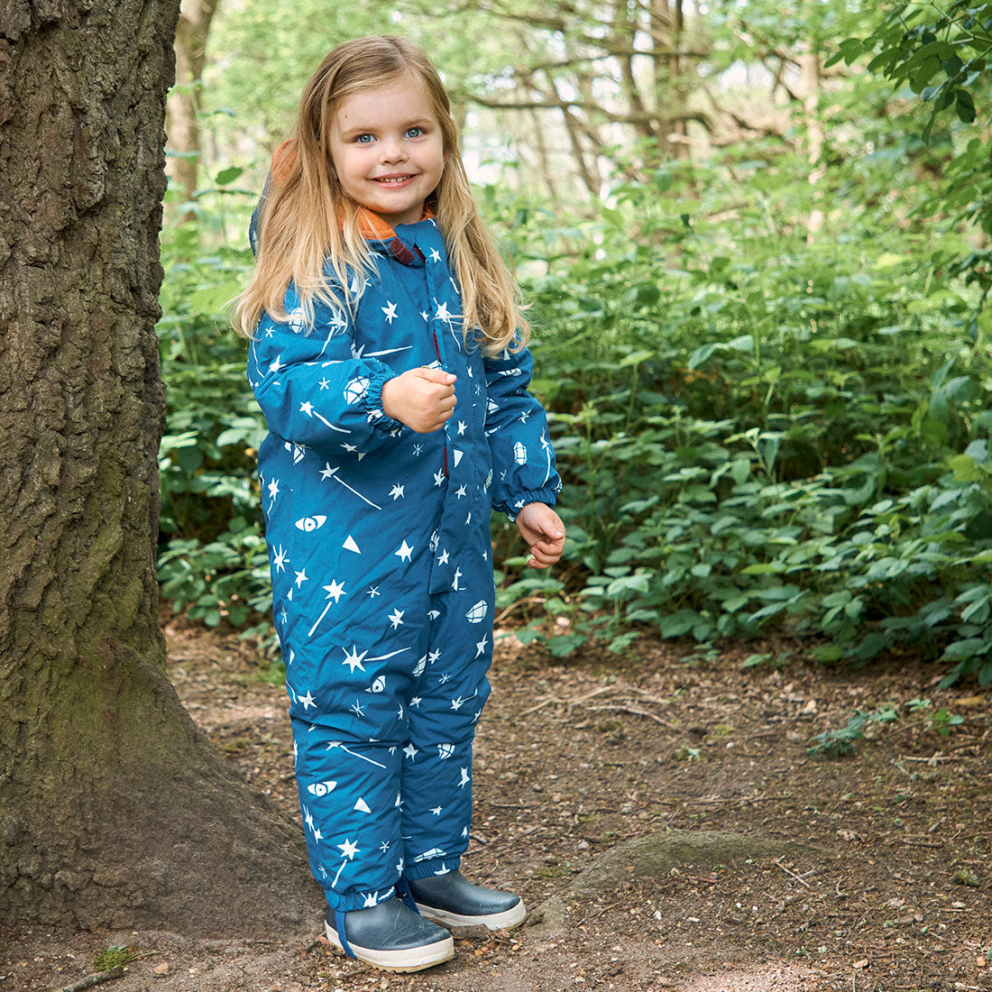A young girl in a forest, wearing a blue 'EcoWarm Waterproof Snowsuit Blue Sky' with star and spaceship patterns, standing next to a tree.
