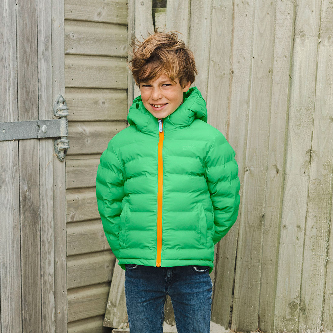 A boy in a Waterproof Puffer Jacket Green smiles, standing in front of a wooden door and fence.