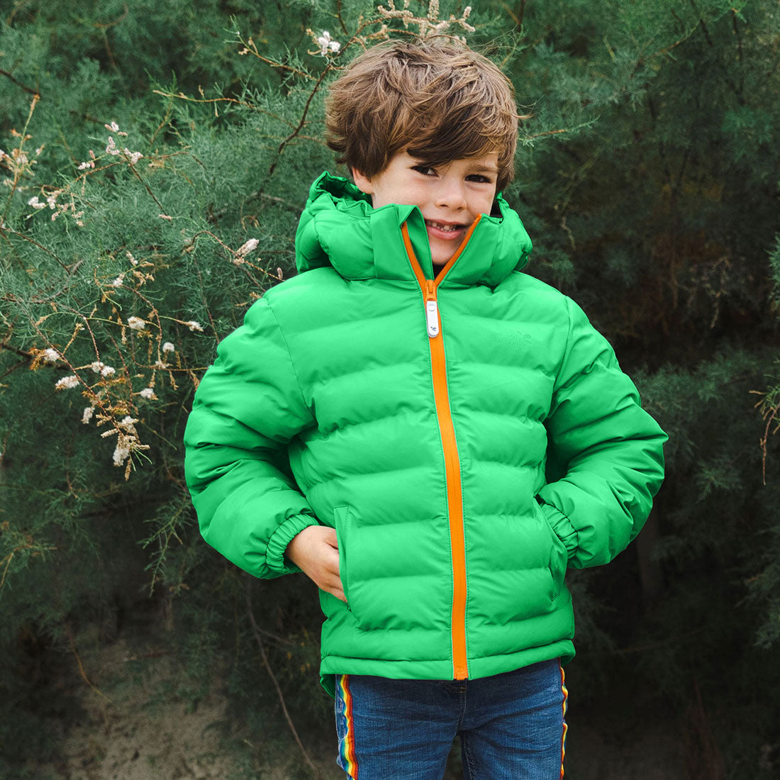 A boy wearing a green waterproof puffer jacket, stands in front of a tree. He has his hands in his pockets, smiling.