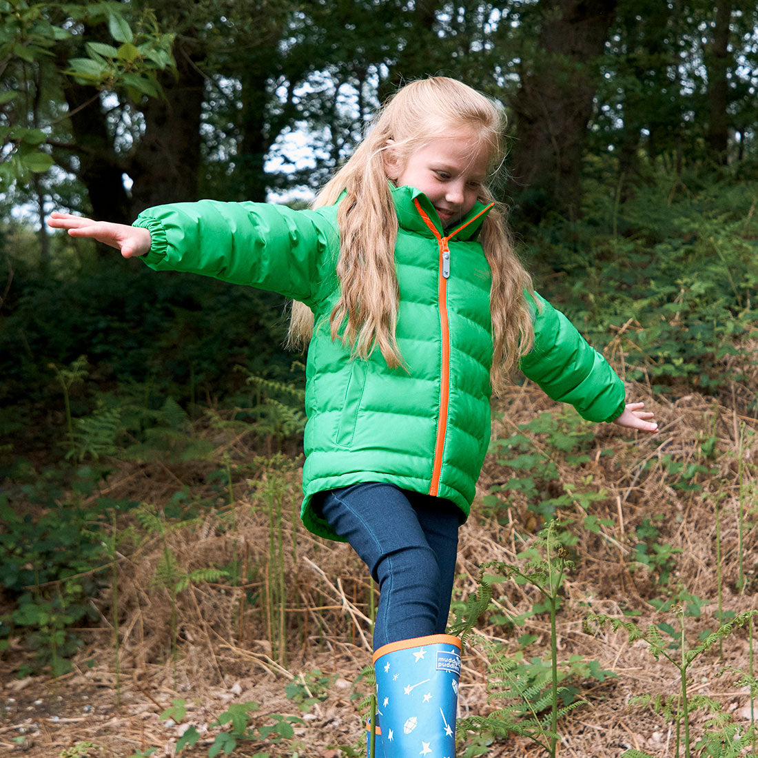 A blonde girl balances in a woodland setting, wearing a green waterproof puffer jacket and blue wellington boots with star designs.