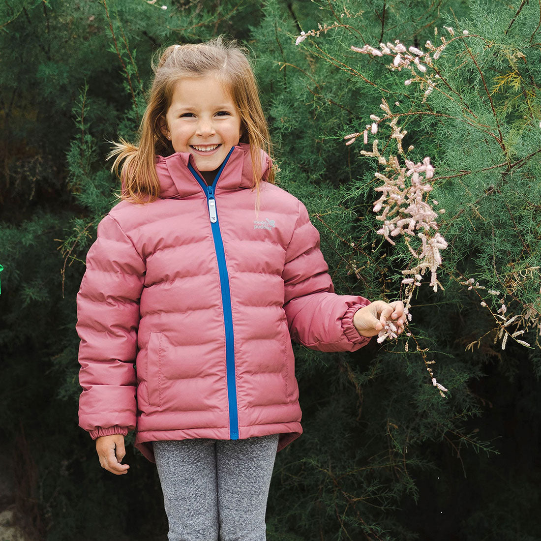 A happy child in a pink puffer jacket with a blue zip, standing in front of green foliage and holding a small branch of pink flowers.