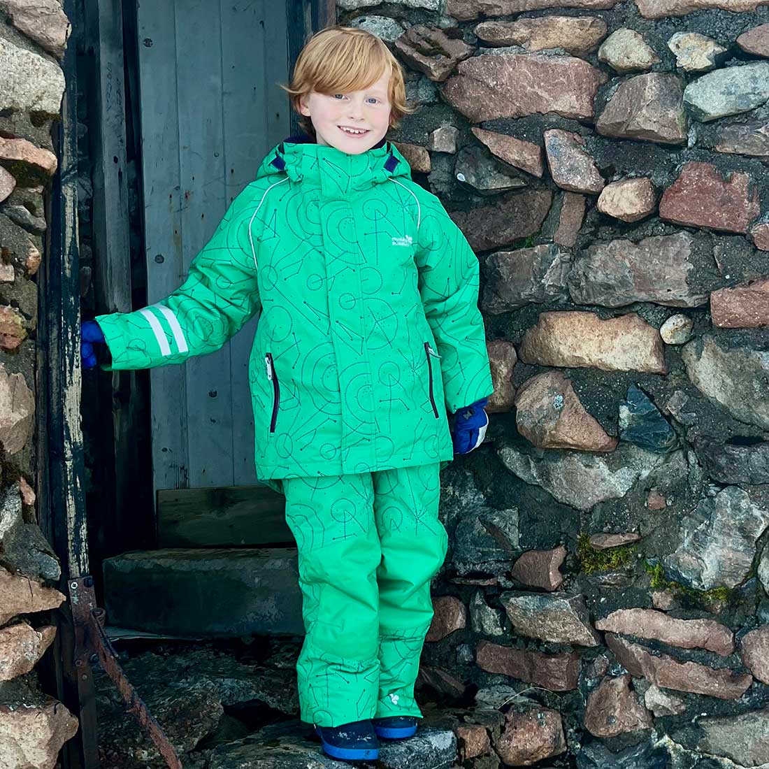 A young boy in a green Blizzard Kids Ski Jacket and matching trousers, stands in a stone doorway with a stone wall backdrop.