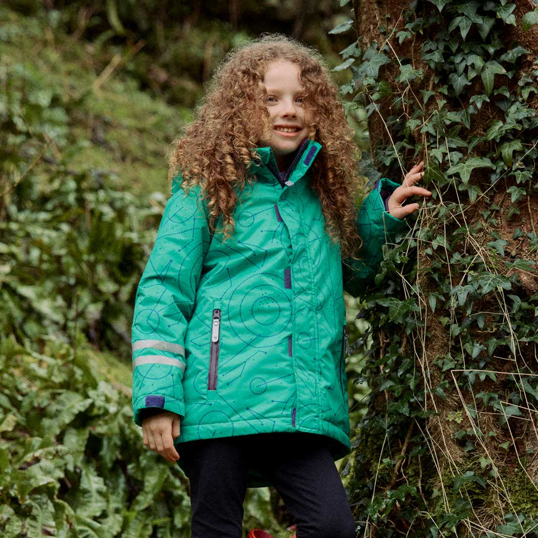 A girl with curly hair wearing a green Blizzard Kids Ski Jacket next to a tree covered in ivy in a wooded area.