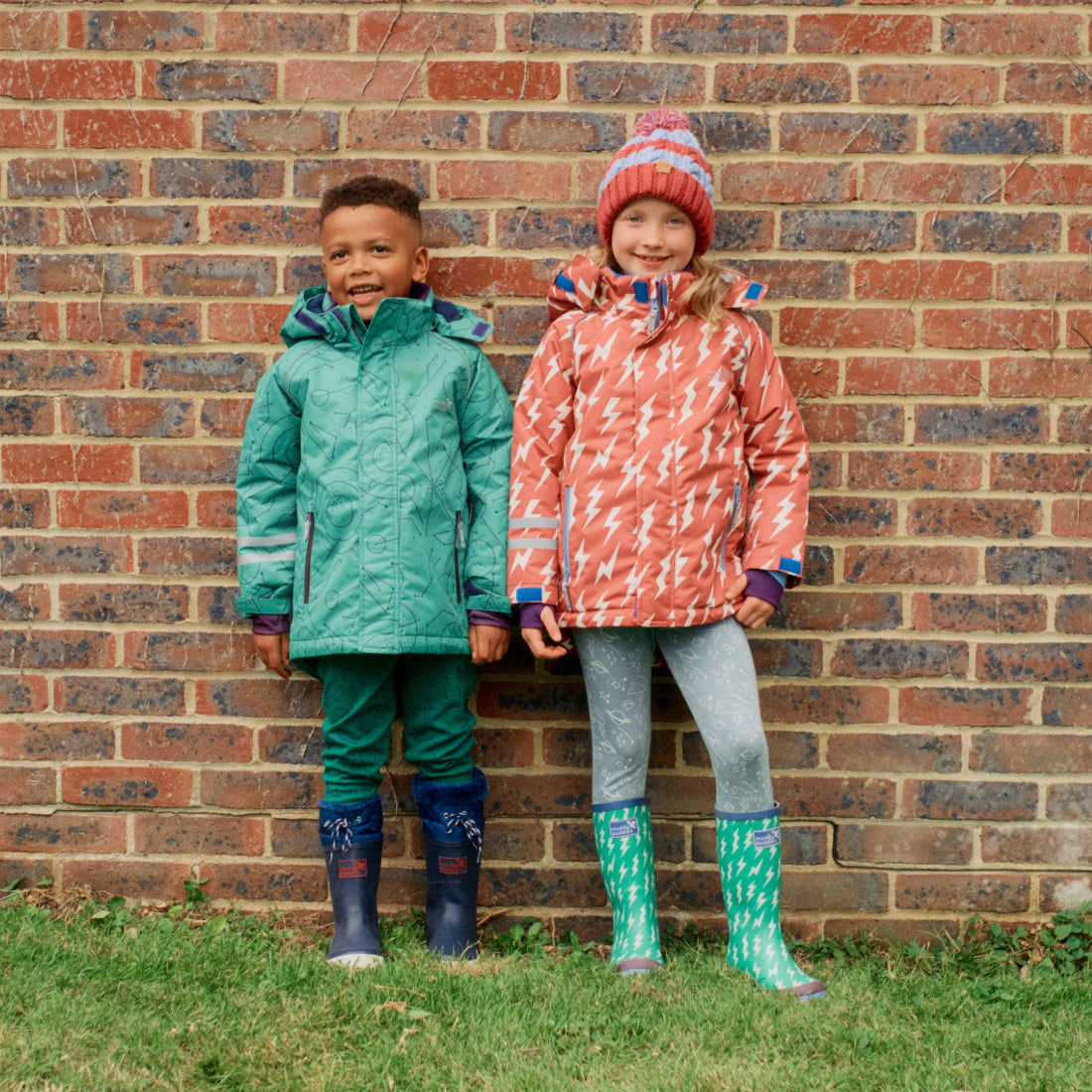Two smiling children stand in front of a brick wall wearing colourful waterproof coats and wellington boots, a Blizzard Kids Ski Jacket Green.