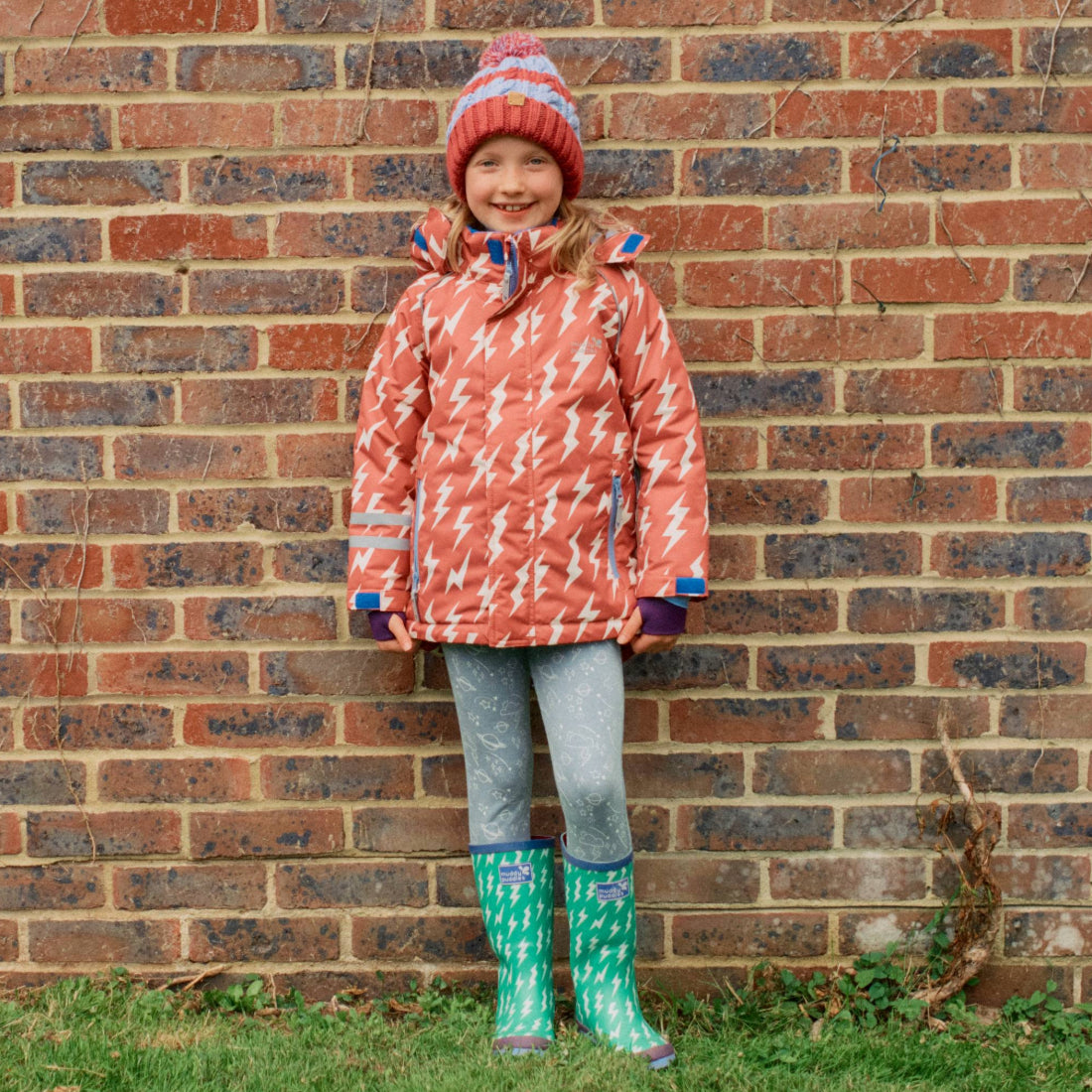 A young girl wearing a Blizzard Kids Ski Jacket Pink, leggings, and wellington boots, standing in front of a brick wall and grass.