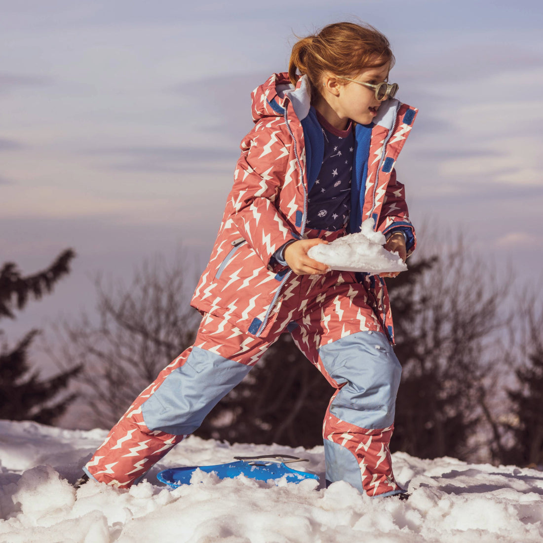 A young girl in a Blizzard Kids Ski Jacket Pink, holding snow outdoors. She's wearing sunglasses and a patterned ski suit.