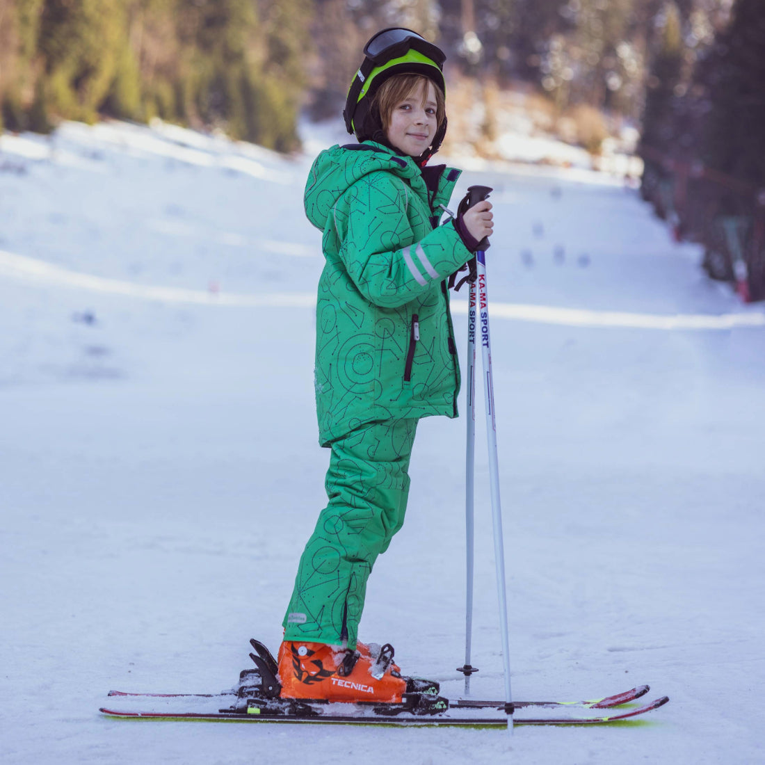 A child in a green Blizzard Ski Salopettes on a snowy slope, wearing a helmet and holding ski poles.