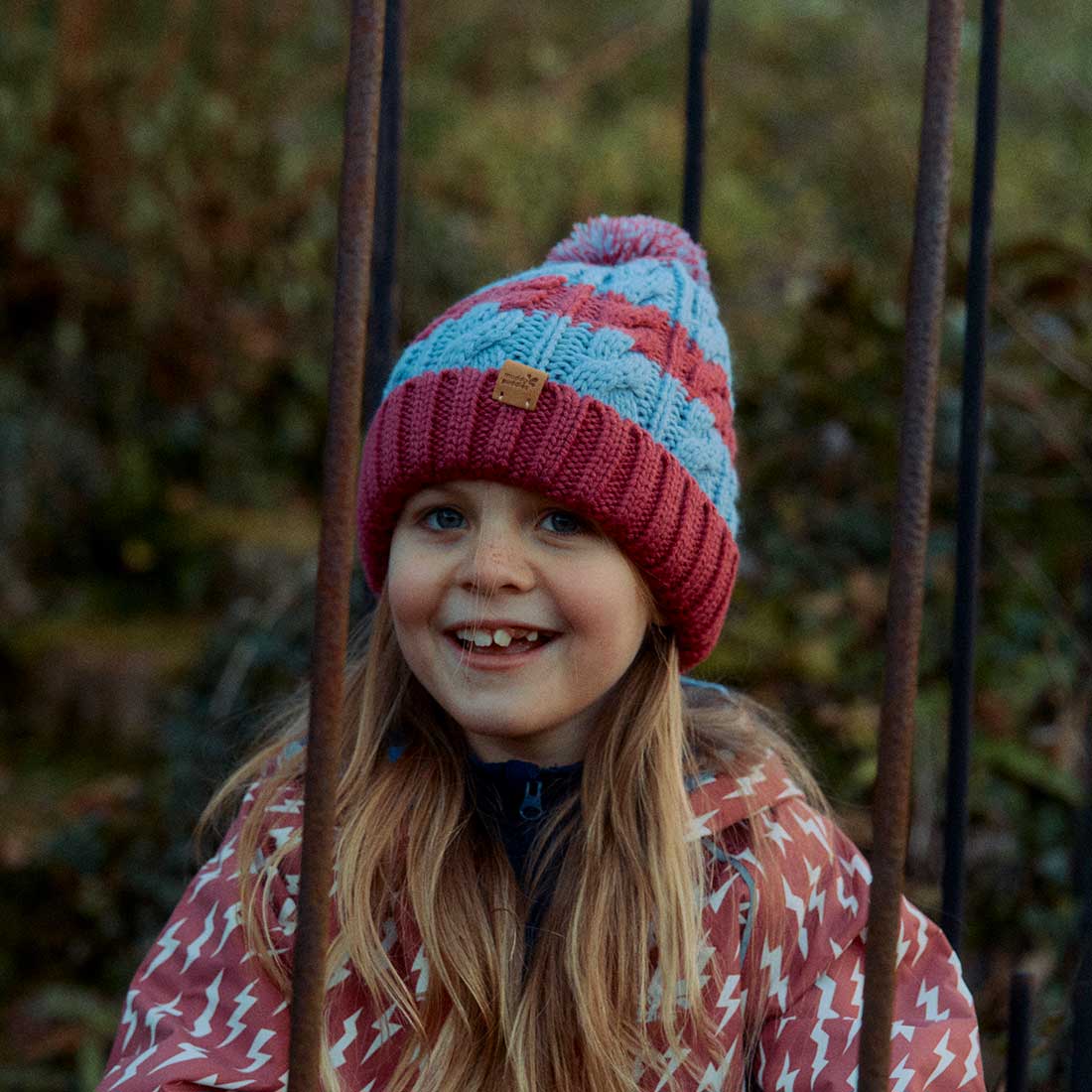A smiling girl wearing a Striped Cable Bobble Hat in pink and blue, standing outside with railings in the foreground.