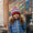 A young girl smiles wearing a Striped Cable Bobble Hat Pink and blue puffer jacket, standing in front of a glasshouse door.