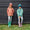 Two children wearing hats and patterned raincoats, standing against a black wooden wall in green boots and trousers in front of grass.