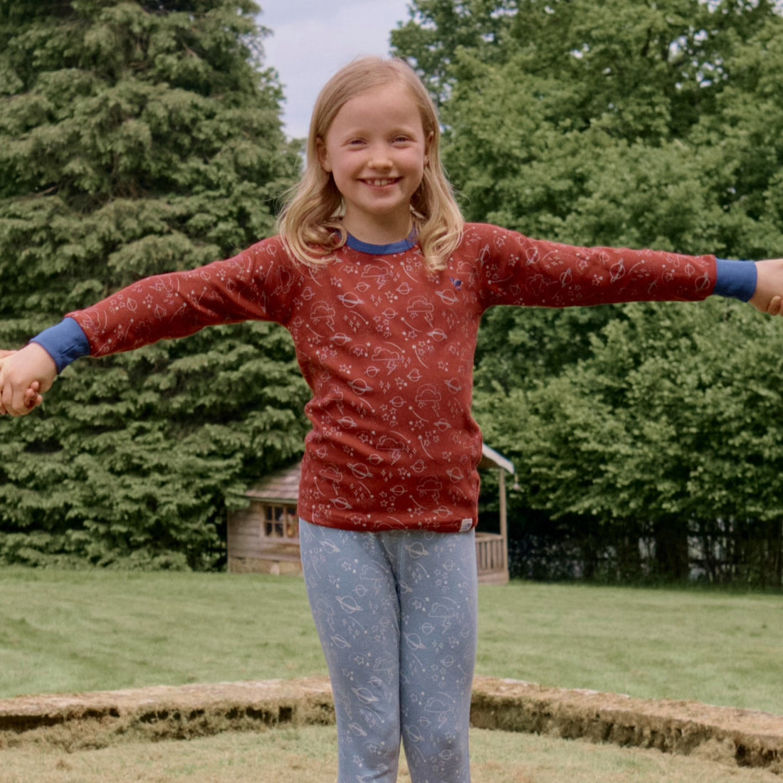 A smiling child wearing a Kids Thermal Top Red, stands with arms outstretched in a grassy outdoor setting with trees and a small wooden structure.