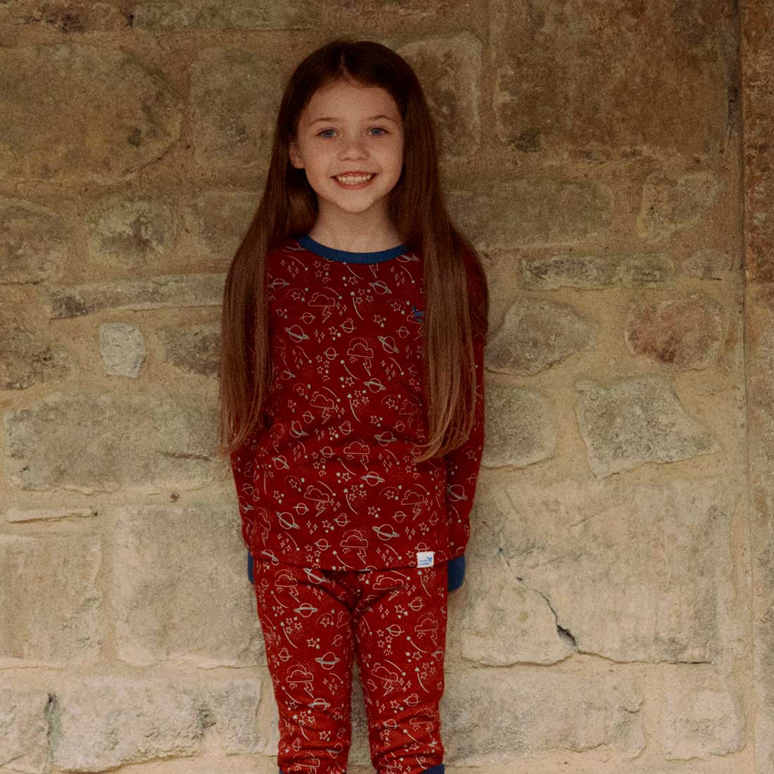 A young girl smiles wearing a red Kids Thermal Top Red pyjama set, stood against a stone wall backdrop.
