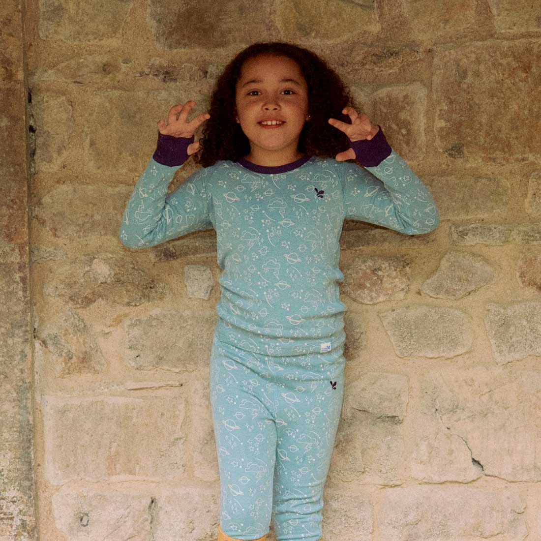 A child in front of a stone wall wearing the Kids Thermal Top Green Sage. They are posing with their hands up.