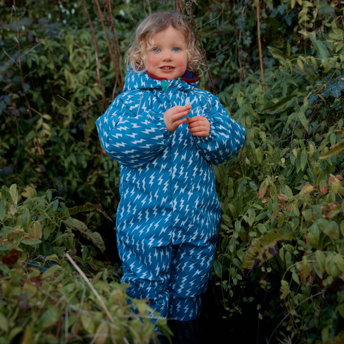 A young child in a blue EcoWarm Waterproof Snowsuit with lightning bolts, standing amidst green foliage.