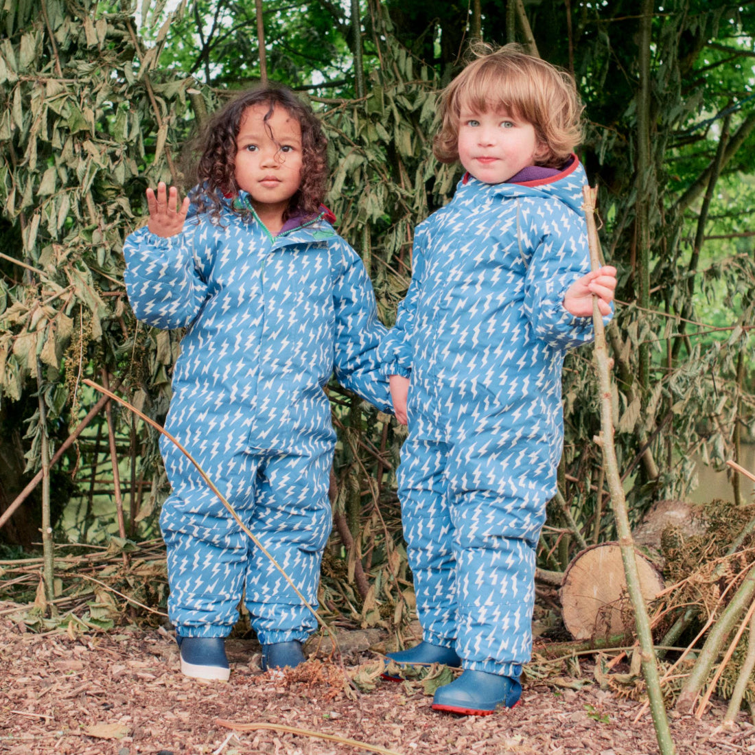 Two children wearing EcoWarm Waterproof Snowsuit Blue, standing in a woodland area, holding hands in their wellington boots.