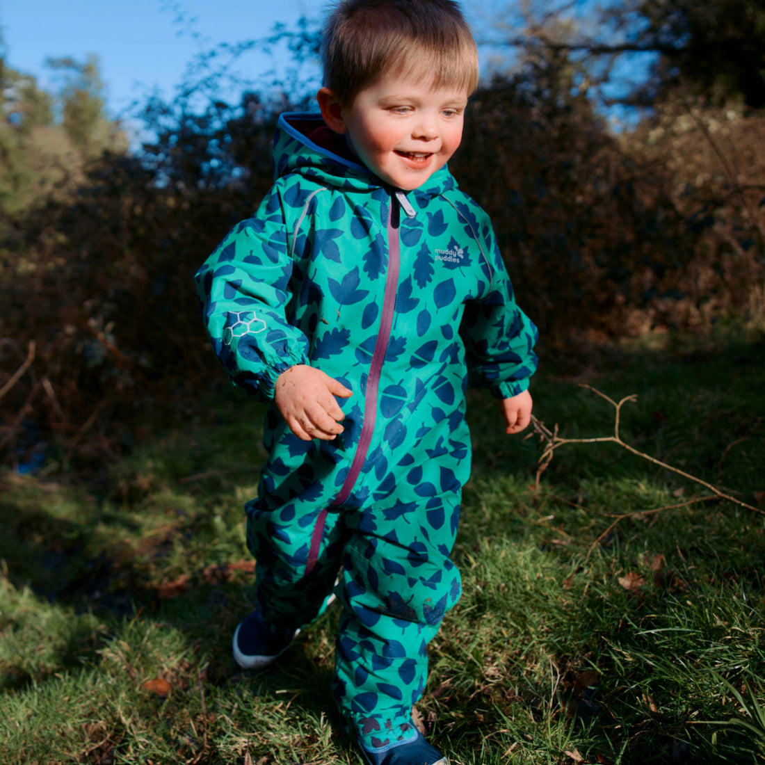 A toddler in an EcoSplash puddle suit, green with a leaf and acorn pattern, smiles as they walk through a grassy outdoor area.