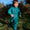 A toddler in an EcoSplash puddle suit, green with a leaf and acorn pattern, smiles as they walk through a grassy outdoor area.