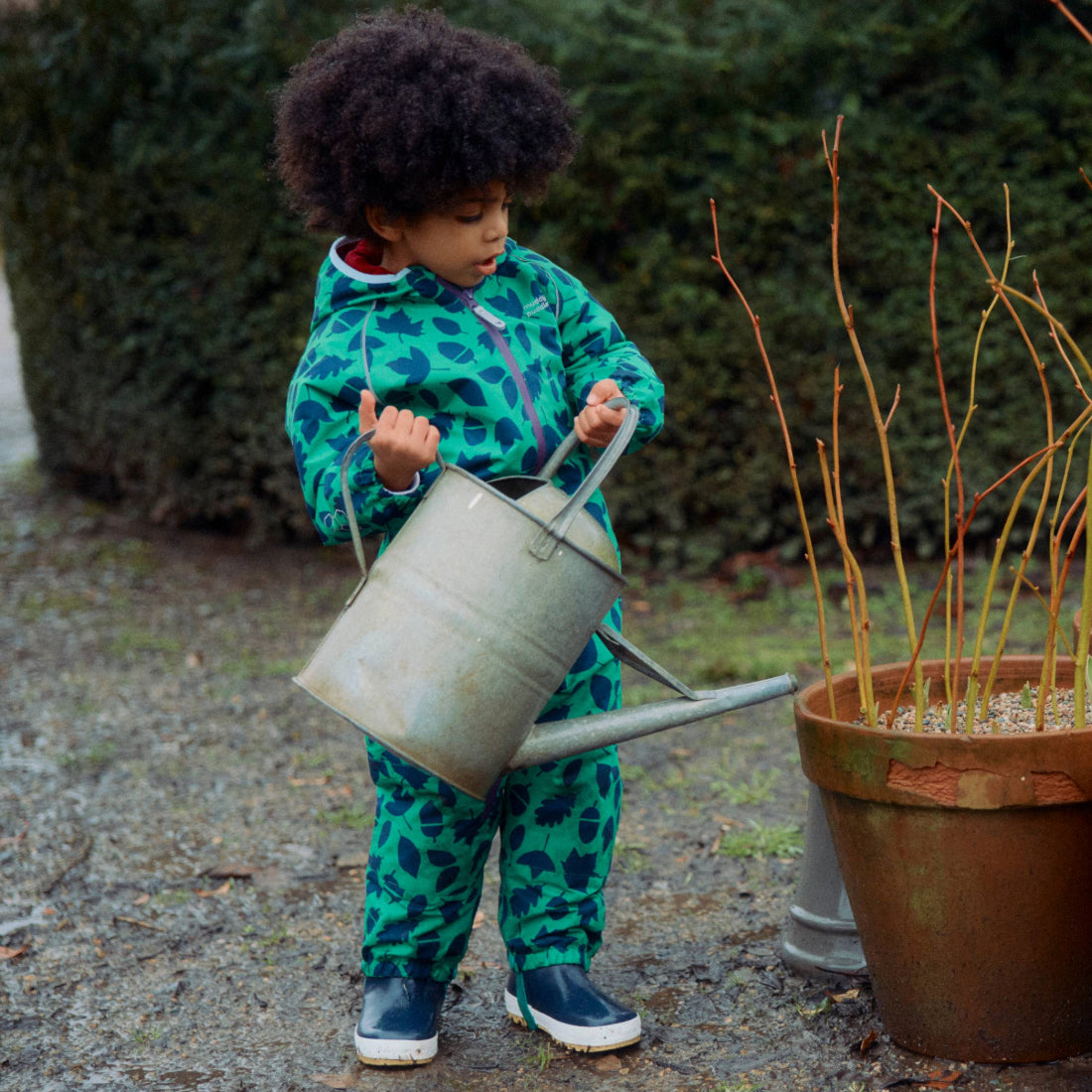 A young child wears an EcoSplash Waterproof Fleece Lined Puddle Suit Green and waters plants in a pot with a watering can, outdoors.