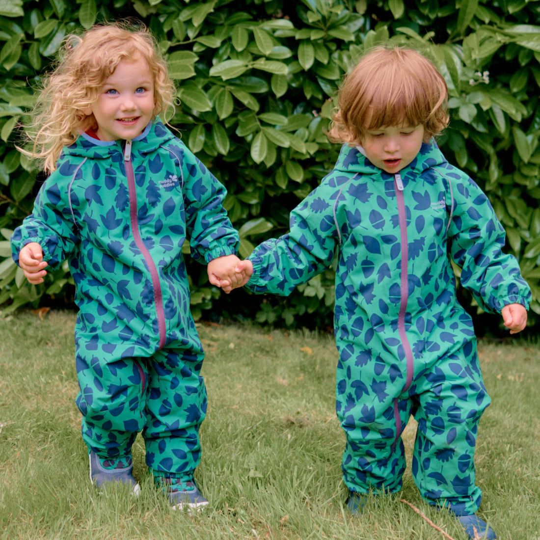 Two young children wearing green EcoSplash Waterproof Fleece Lined Puddle Suits, holding hands on a grassy lawn, with a green bush behind.