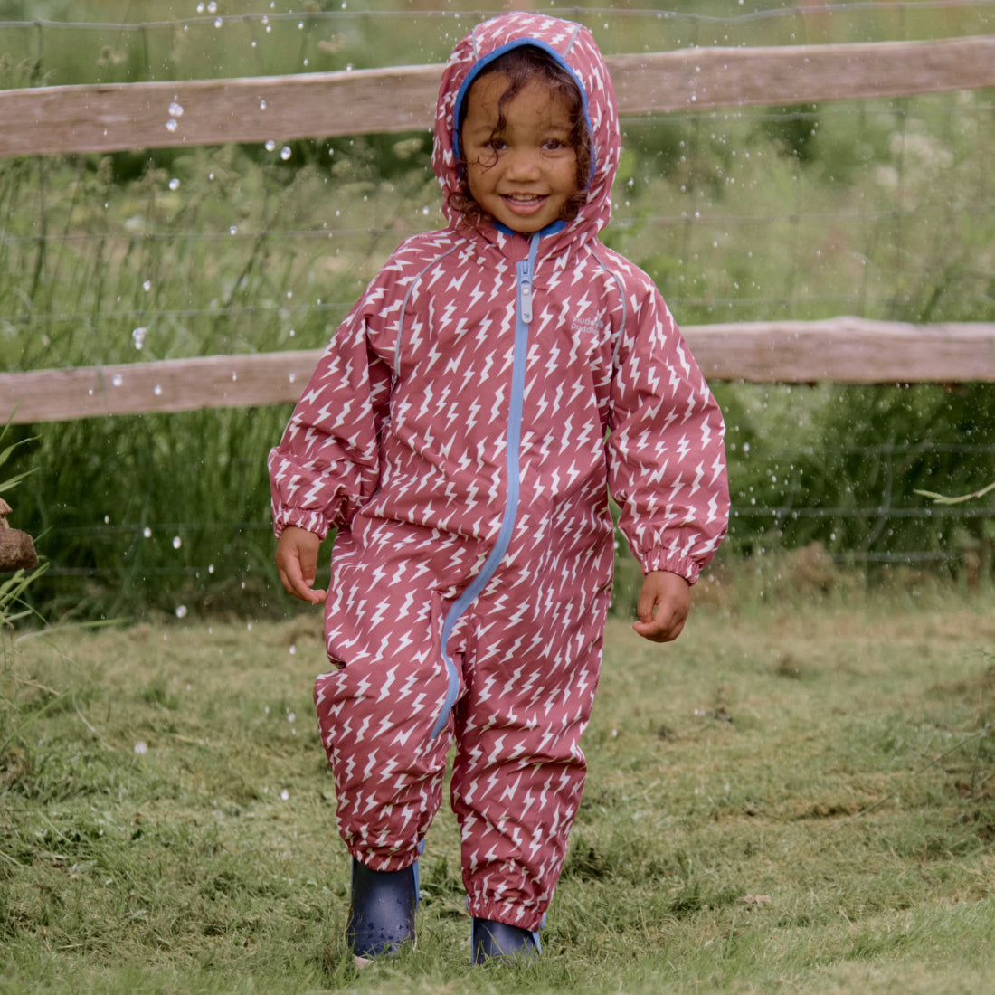 A smiling toddler in an EcoSplash Waterproof Fleece Lined Puddle Suit Pink, standing in a garden with a wooden fence behind.