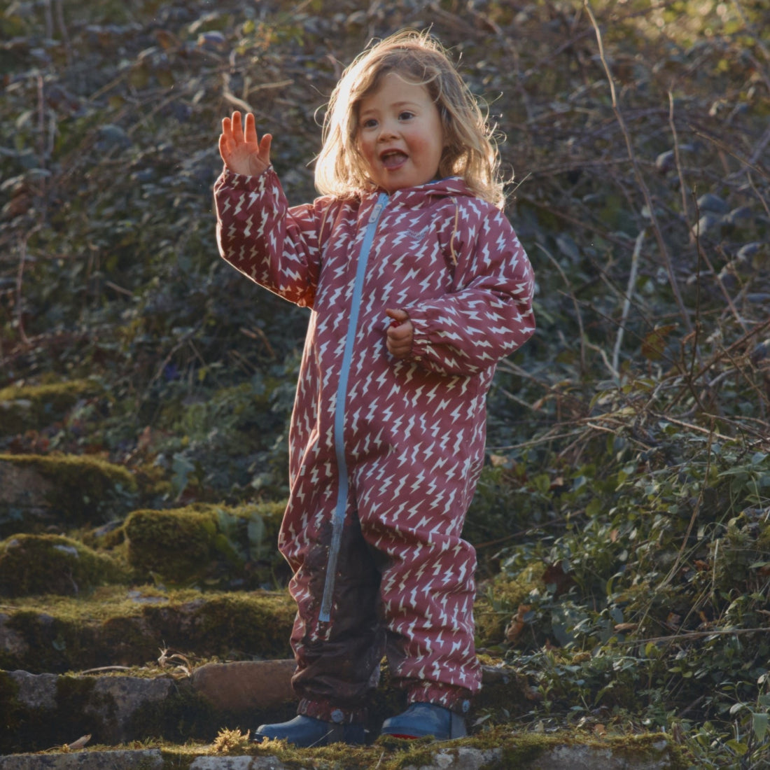 A young child in an EcoSplash Puddle Suit waves with a grin. The suit is pink with white lightning bolts, in an outdoor setting.