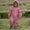 A smiling toddler in an EcoSplash Waterproof Fleece Lined Puddle Suit Pink, standing in a garden with a wooden fence behind.