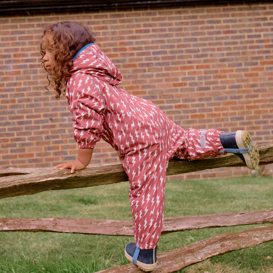 A child in an EcoSplash Waterproof Fleece Lined Puddle Suit Pink climbing a wooden fence against a brick wall backdrop.