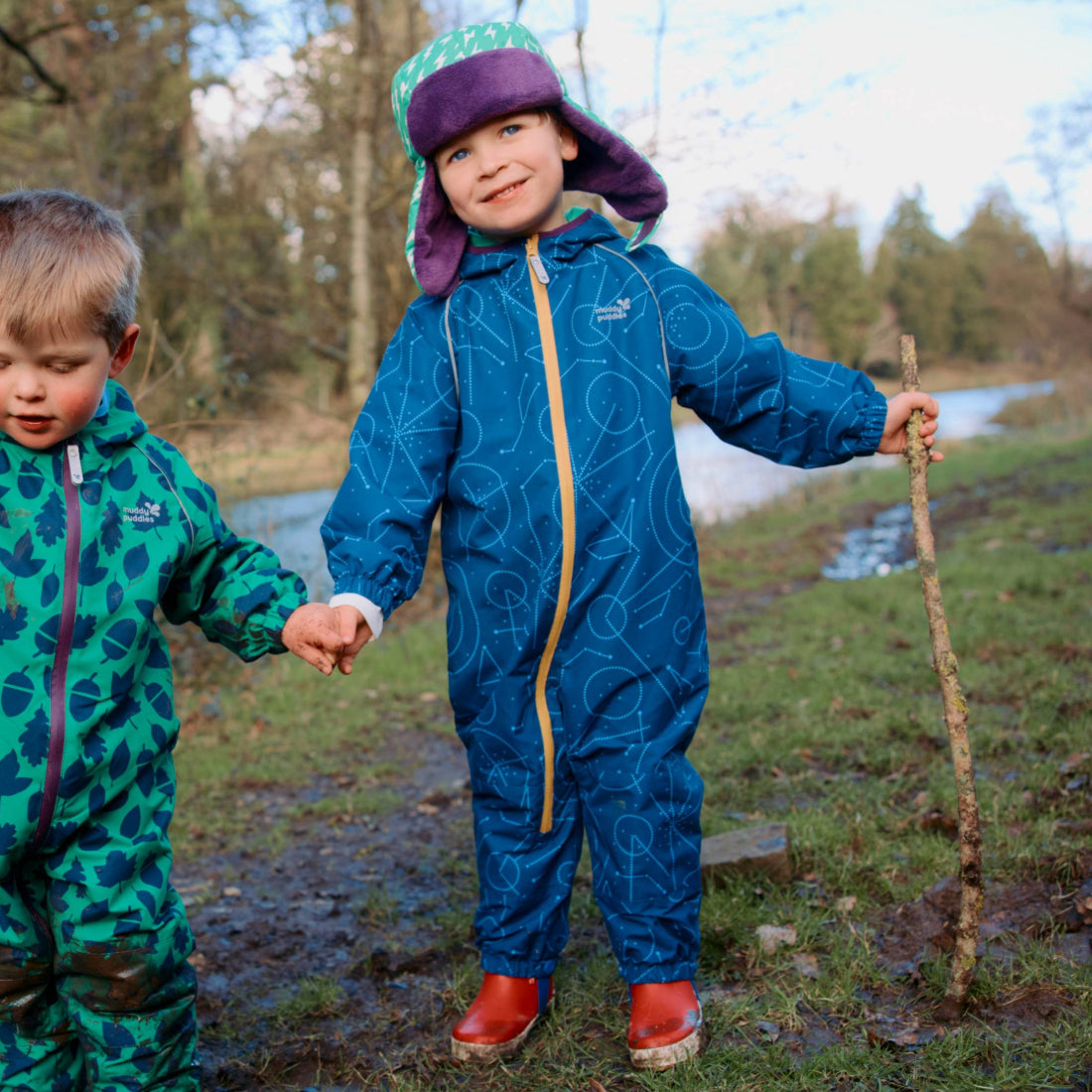 Two children wearing Muddy Puddles EcoSplash Puddle Suits, one blue, one green, standing by a river in a muddy park.