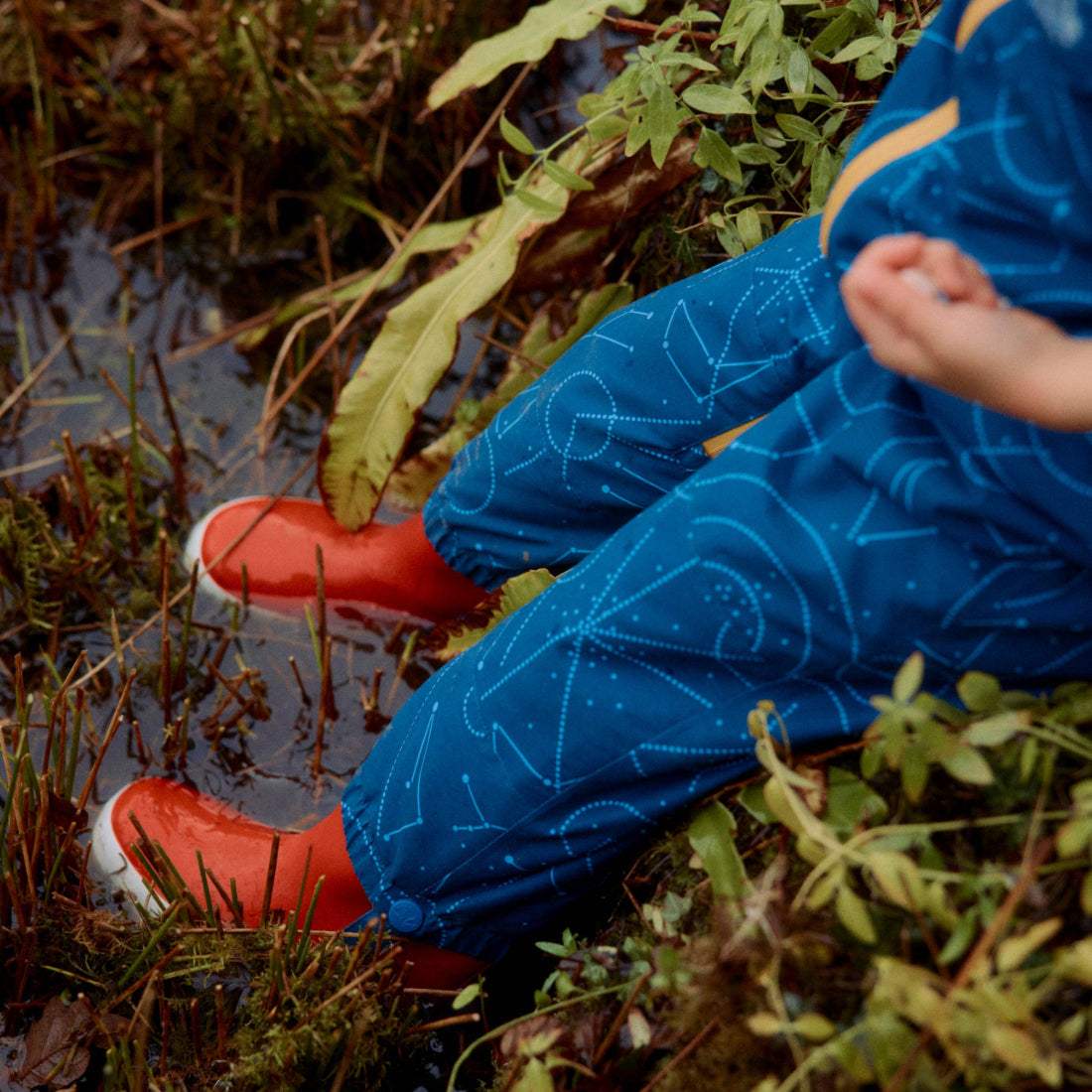 EcoSplash Waterproof Fleece Lined Puddle Suit Navy: A child in a blue constellation-patterned puddle suit and orange wellies sits by a puddle.