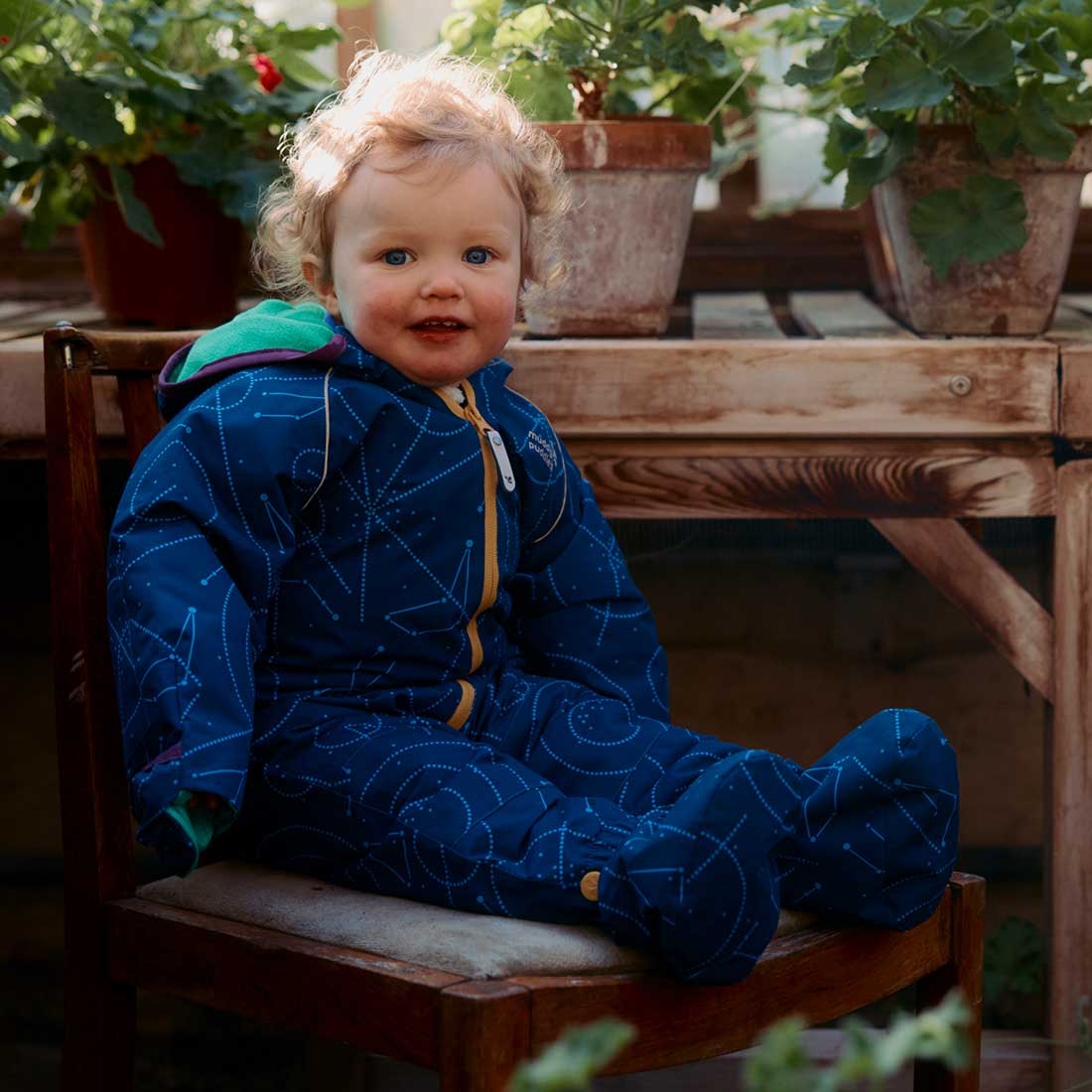 A baby wearing a navy EcoSplash Waterproof Fleece Lined Puddle Suit, seated on a chair in a greenhouse surrounded by potted plants.