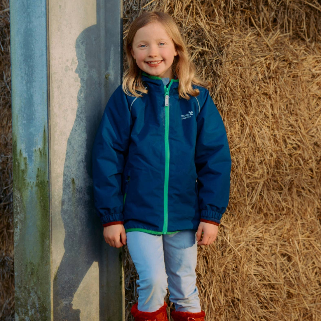 A young girl smiles wearing a 'EcoSplash Waterproof Fleece Lined Jacket Navy', next to a concrete pillar and straw stack.
