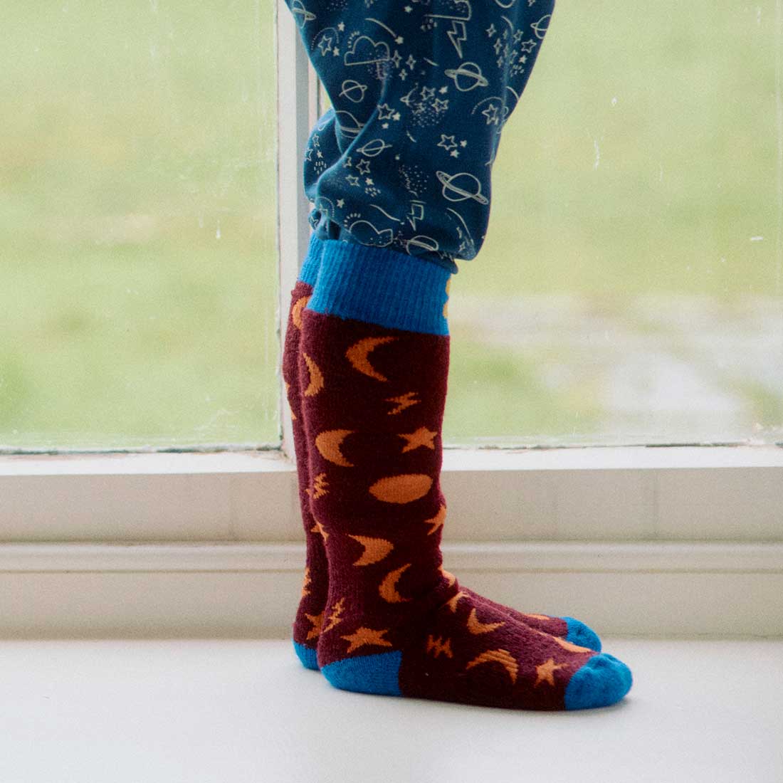 Puddlestomper Welly Socks Purple, knee-high socks with a blue toe and cuff, featuring yellow stars, moons, and planets on a maroon background.