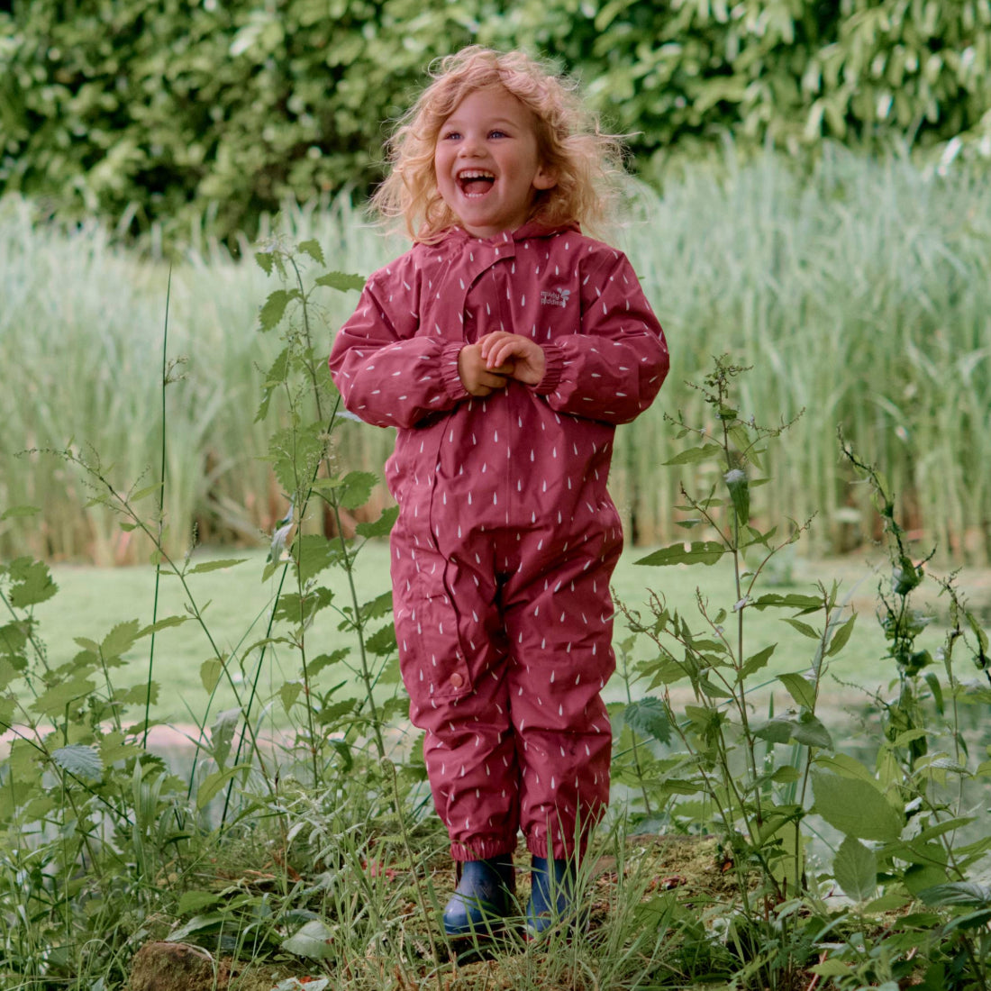 A happy child in a patterned maroon jumpsuit stands amidst greenery, wearing wellies and laughing with hands clasped.