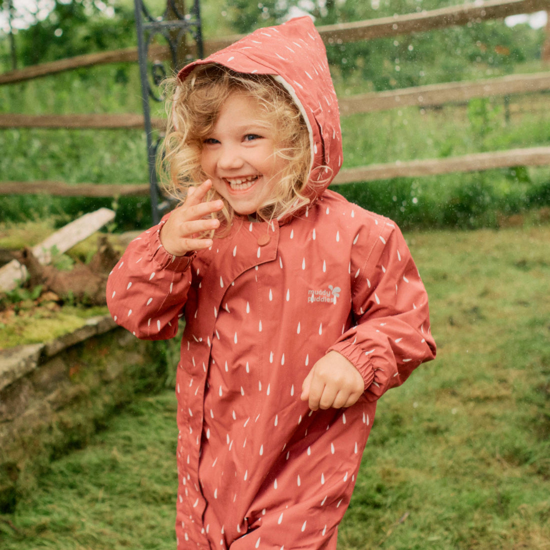 A happy child in a pink 3 in 1 Waterproof Scampsuit patterned with raindrops stands outdoors in the rain, grinning widely.