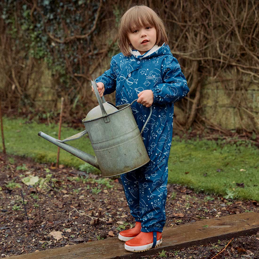 A child in a navy 3 in 1 Waterproof Scampsuit, patterned with planets, stars & clouds, stands on wood holding a watering can, in a garden.